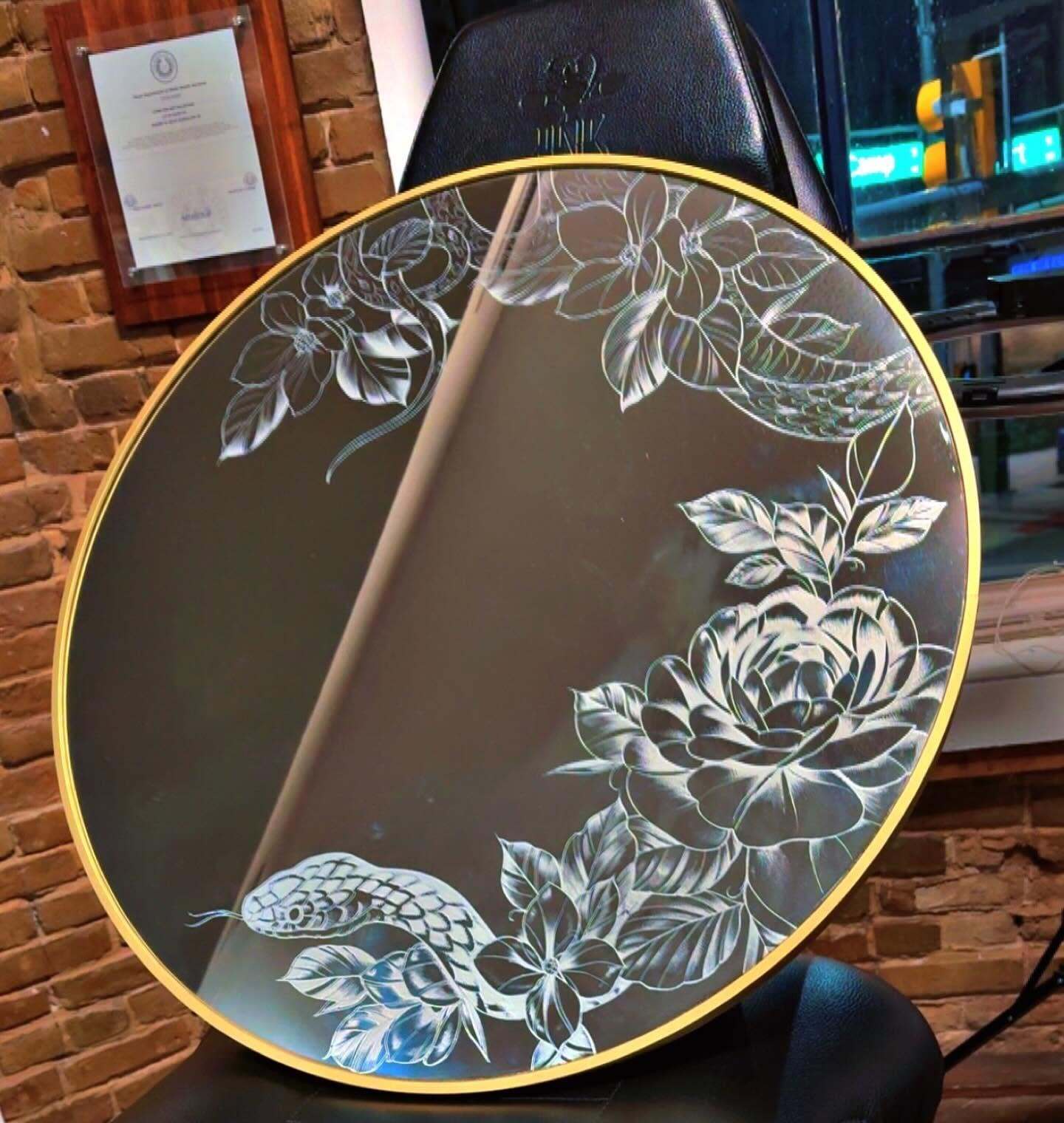 A round gold-framed mirror hand-etched with roses, leaves and a snake, resting on a tattoo chair.