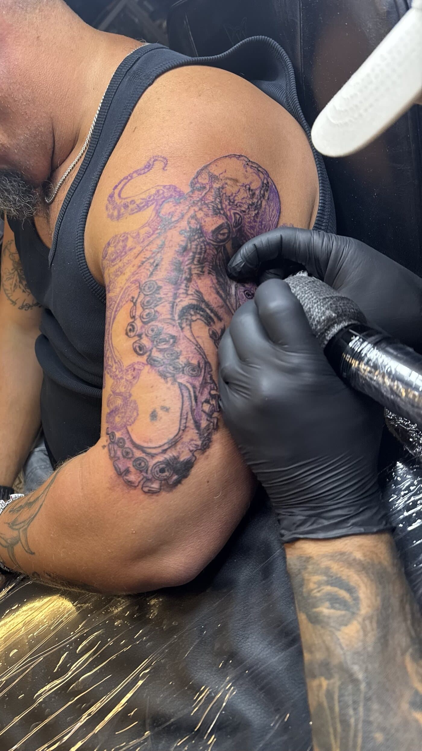 An octopus tattoo in progress on an upper arm: purple stencil lines with the first black linework going in, Jeremiah’s gloved hands at work.